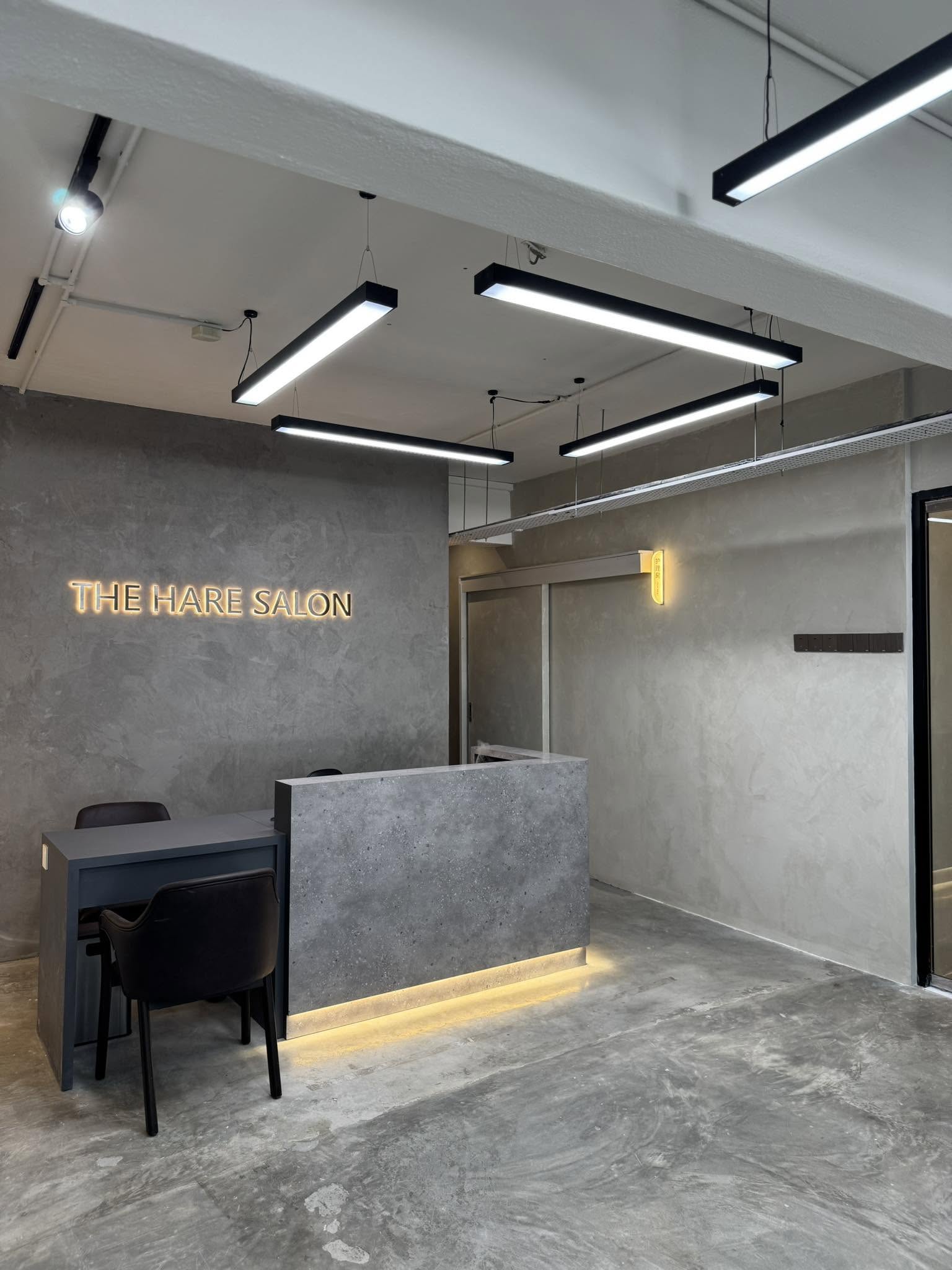 Inside The Hare Salon — a calm, micro-cement studio with black linear pendant lights and the gold ‘THE HARE SALON’ sign glowing over a concrete reception desk